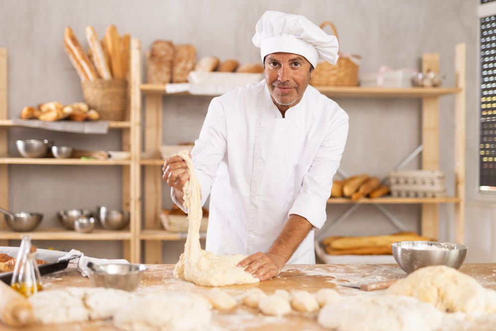 Male baker stands at work bench, kneading and shaping dough to make bread, croissants and baguettes