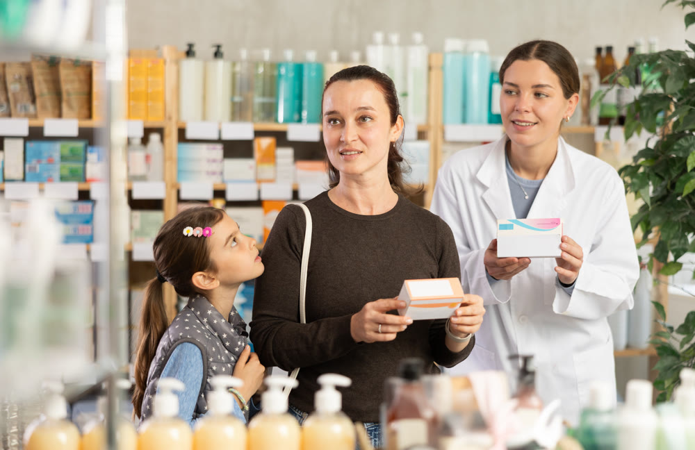 Woman with schoolgirl daughter are busy choosing tablet box at pharmacy, pharmacist in background