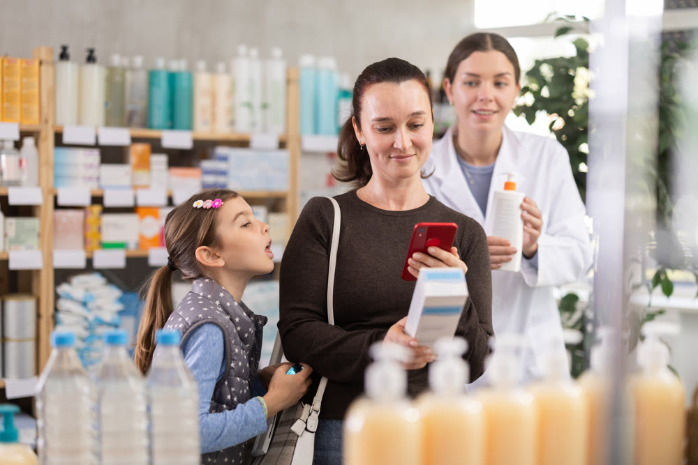 Woman with daughter in pharmacy scans QR code on cream, pharmacist in background