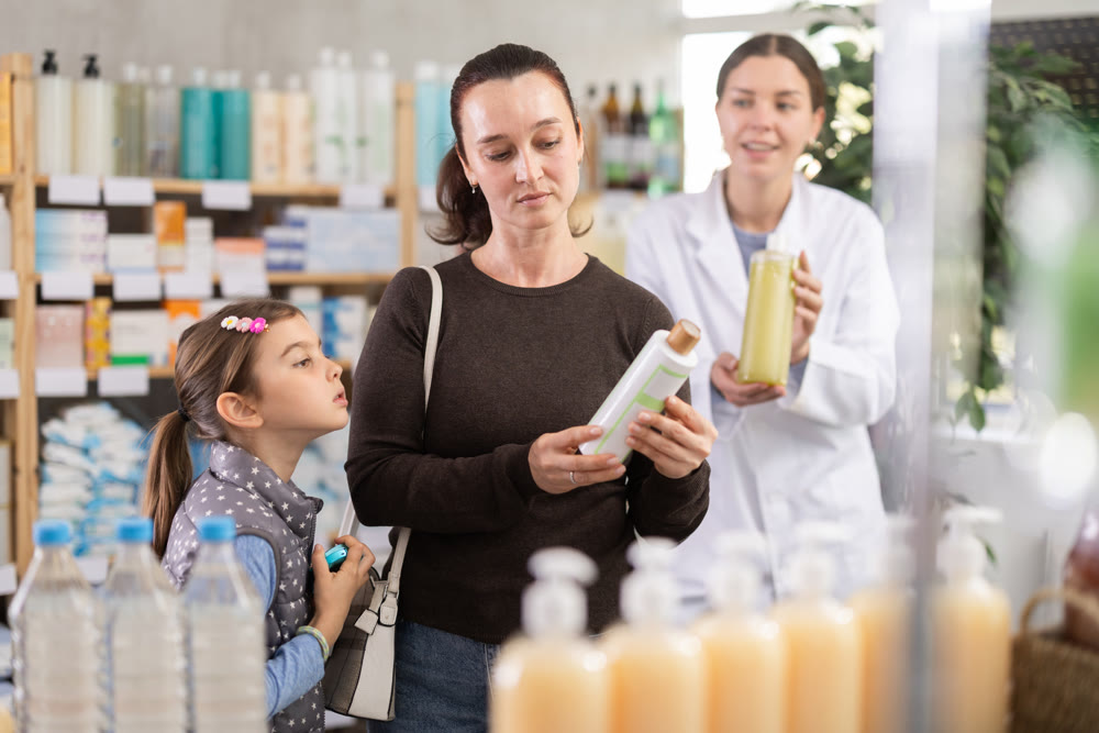 Woman with schoolgirl daughter are busy choosing shampoo at pharmacy. Pharmacist in background