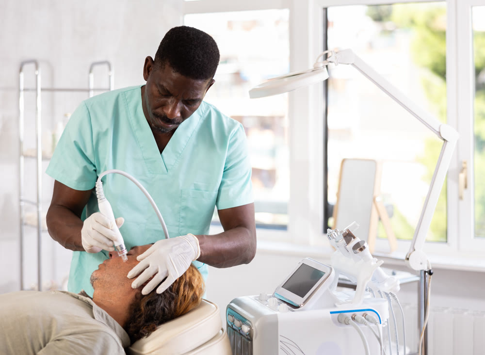 African male cosmetologist performs skin cleansing procedure for man client with beauty machine