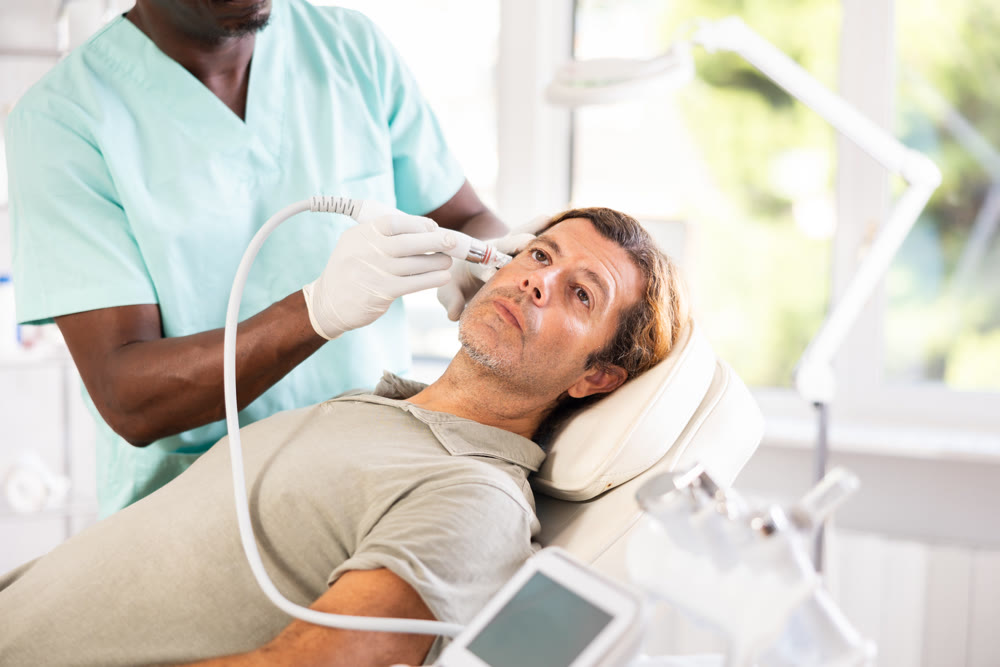 African male cosmetologist performs skin cleansing procedure for man client with beauty machine