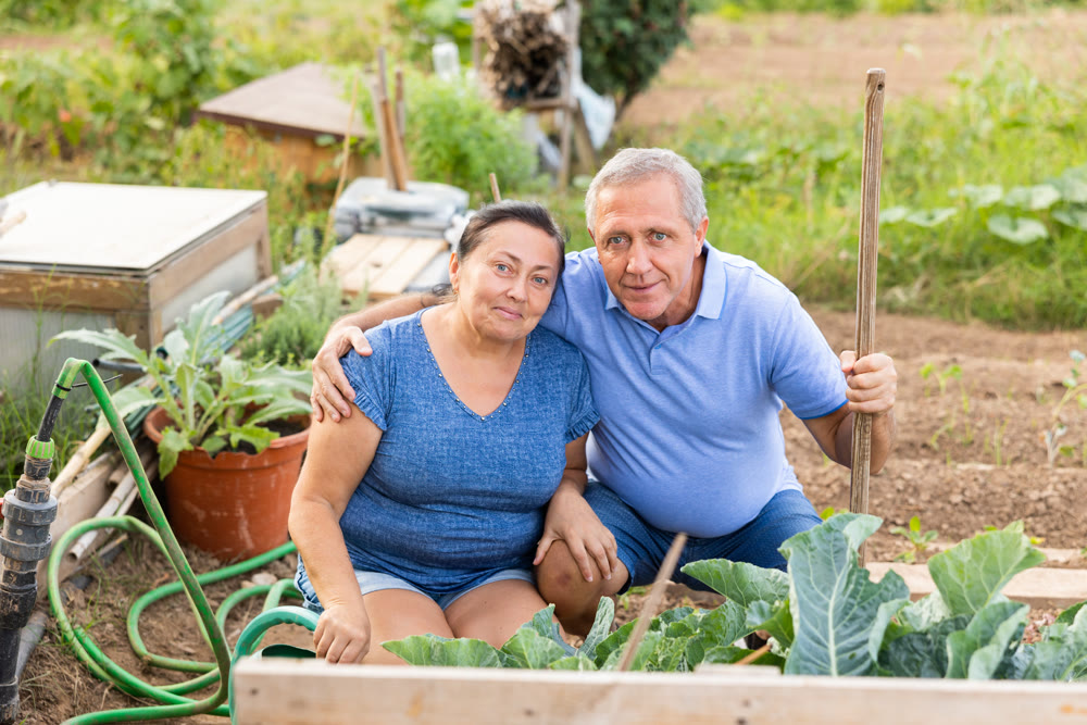 Smiling aged woman and man standing in vegetable garden at smallholding