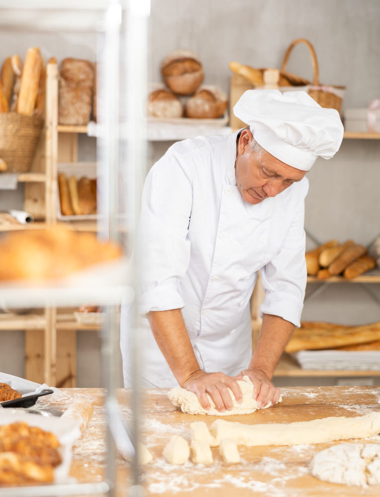 Senior man works in bakery as baker, kneads dough, works with flour.