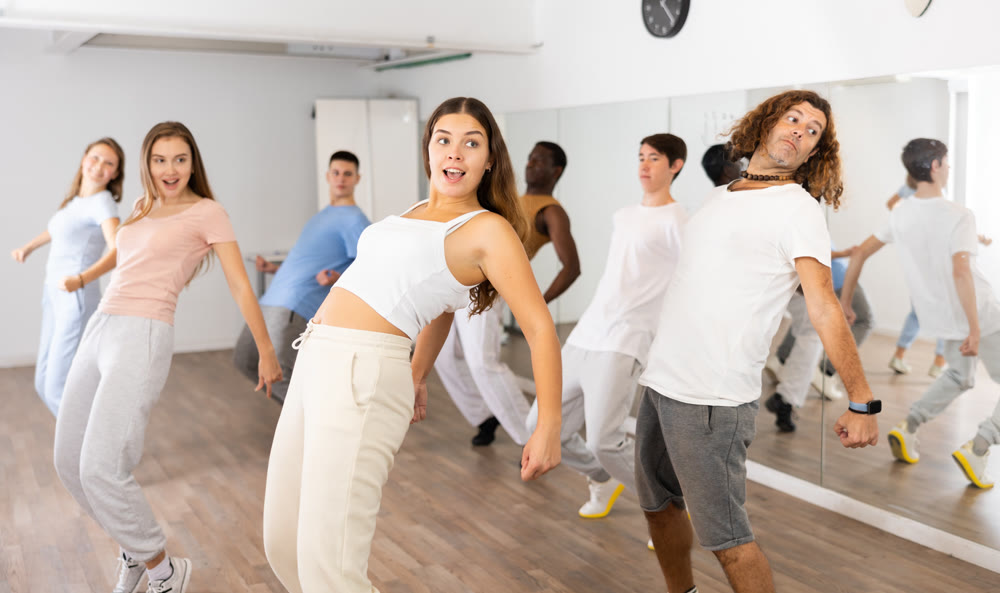 Excited diverse men and women of different ages leaning back while standing on their toes in dancing class for adults