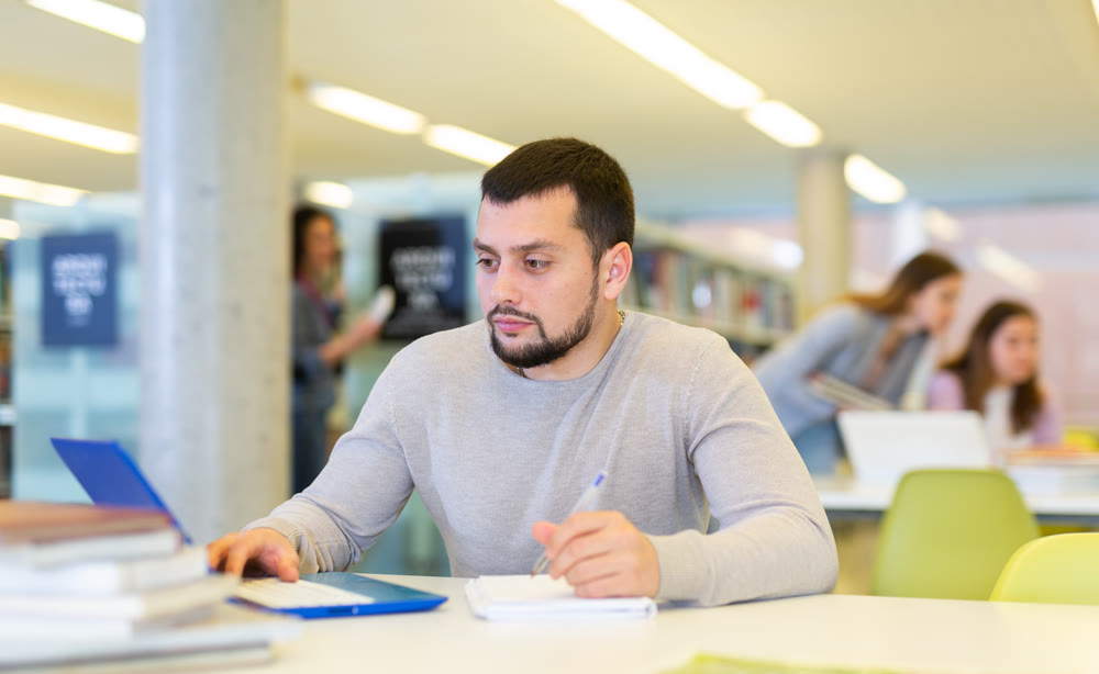 Focused young student studying with books and laptop in library