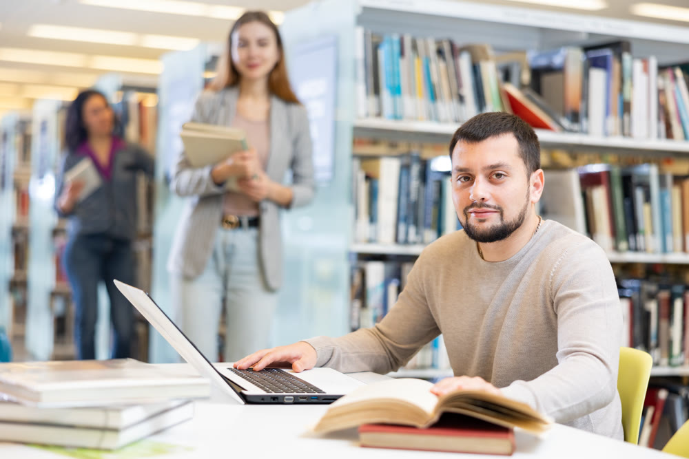 Middle aged man reading books and working on laptop in library