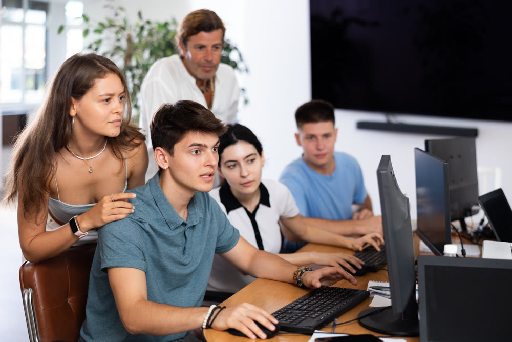 Girl stands behind sitting students, looks at monitor and helps in execution of programing teamwork
