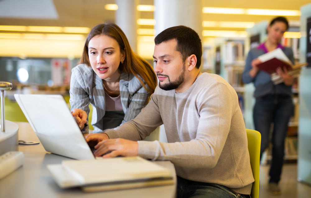 Man and woman spending time together in library, talking, work on laptop