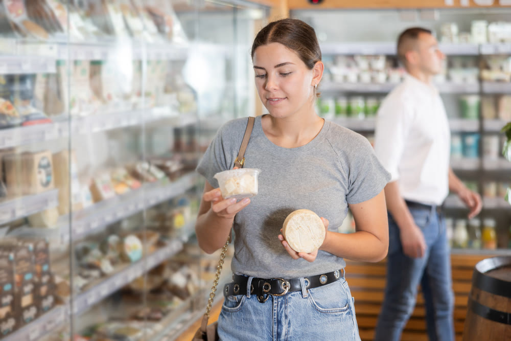 Girl in bio food store holds cheese of different varieties in hands and is thinking about buying