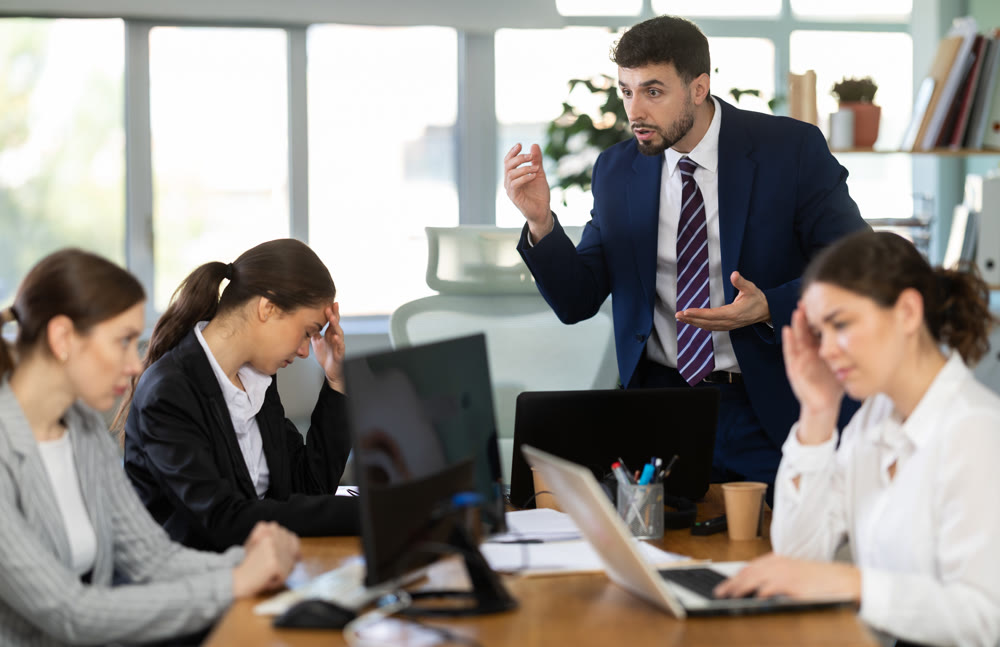 Young man standing and scolding sad seated colleagues in office