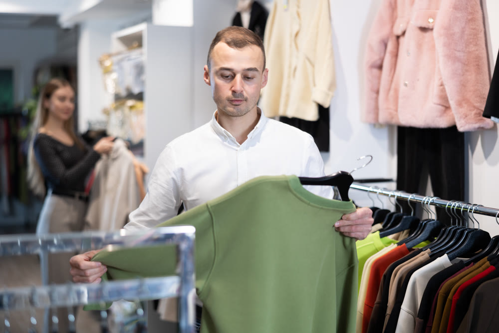 Man stopped near showcase with outerwear, holds blazer hanger in hands and examines product