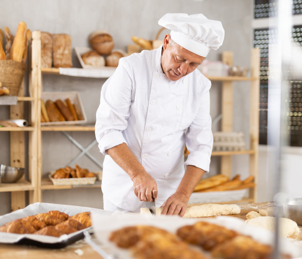 Elderly male baker cutting dough with scraper