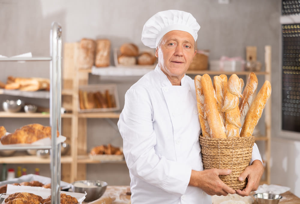 Smiling older baker holding basket of golden baguettes
