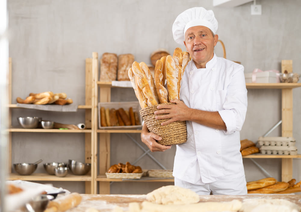 Bakery mature man employee holds basket of ready-made baguettes.