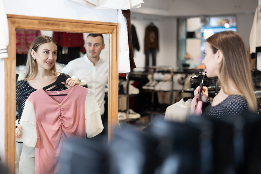 Girl stands in front of mirror hold hanger with blouse, puts it to herself and admires her reflection.