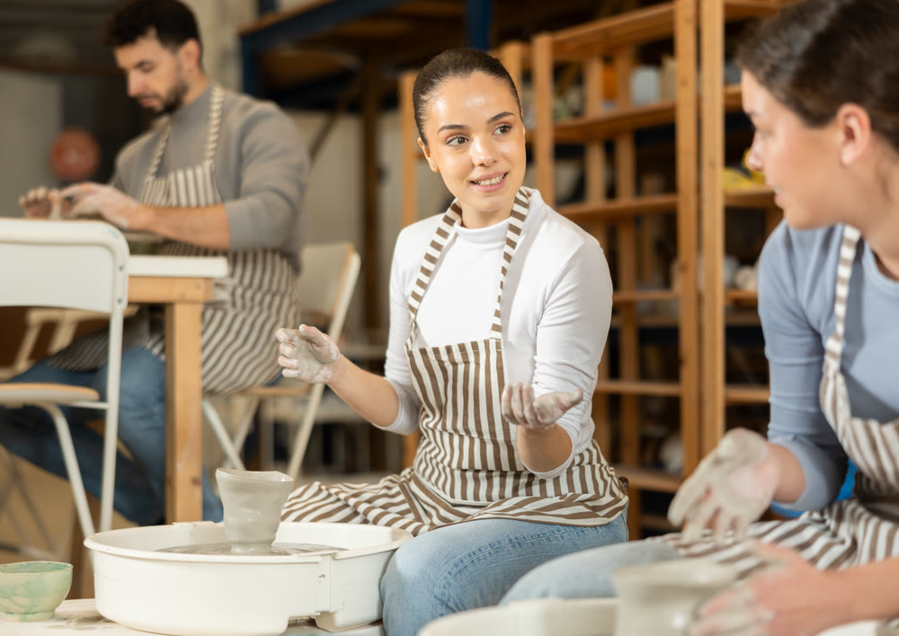Two girls mold products on potters wheel and talk to each other.