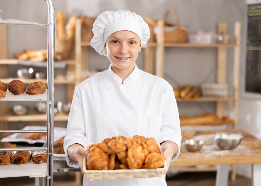 Young female baker apprentice holding tray of baked croissants