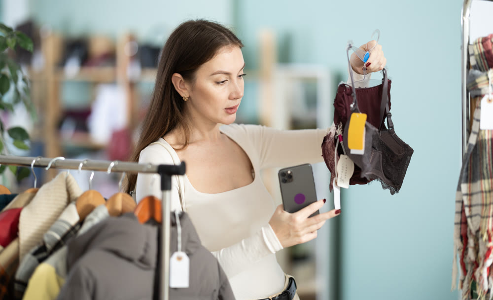 Focused female customer dressing in ordinary clothes holding bras in hands and scanning barcode with smartphone in underwear store