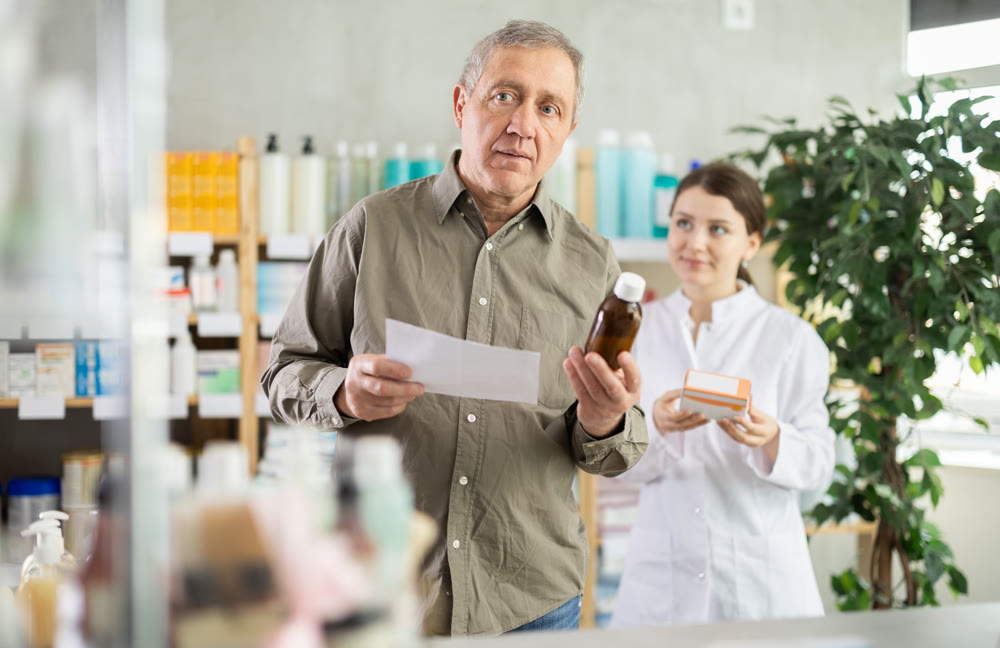 Elderly male customer chooses cough syrup in modern pharmacy. Female pharmacist helps with choosing medicine