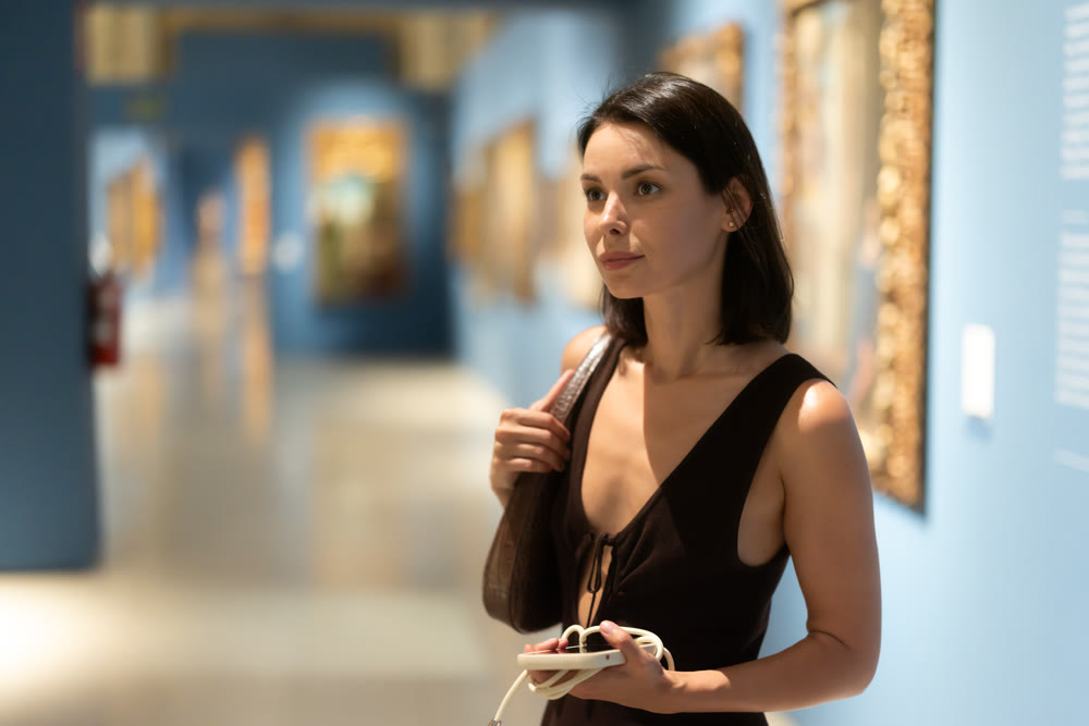 Woman with phone in hands in museum hall, view painting exhibition