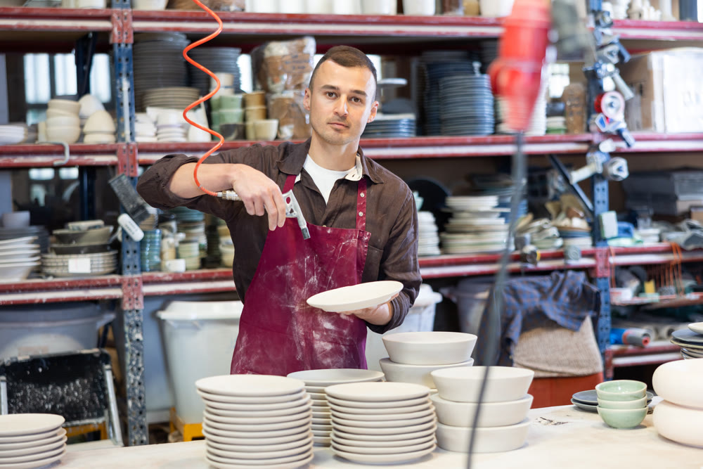 Artisan dusting off ceramic wares with air blow gun in pottery workshop