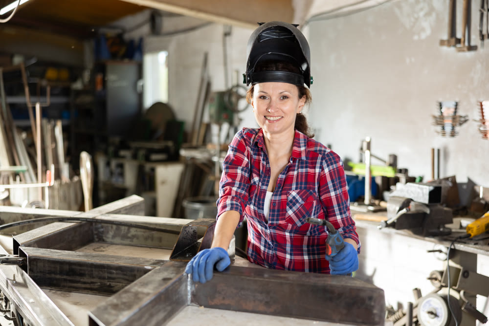 Portrait of a female welder standing with a welding semi-automatic machine and a safety helmet in metal machining workshop