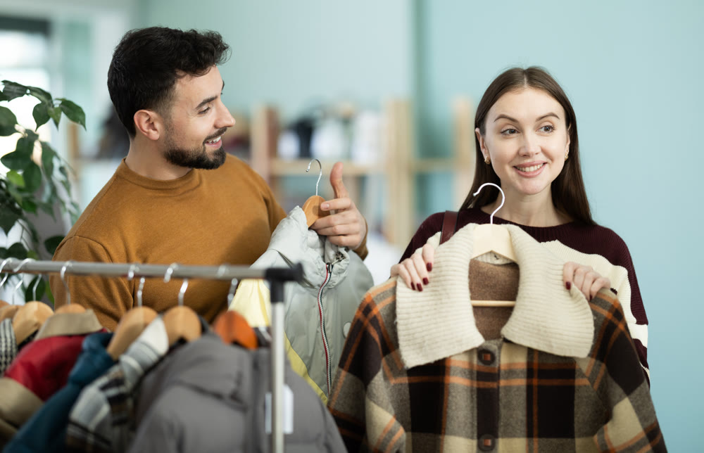 European couple buyers selecting new coat for cold season, woman looking and man helping choose right size in boutique
