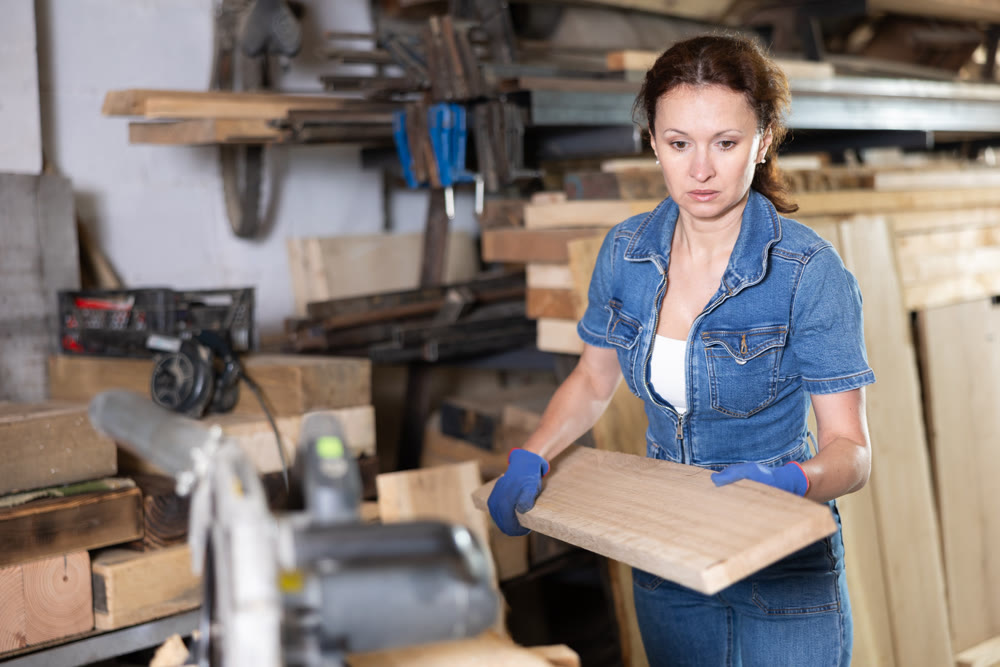 Woman employee of carpentry workshop takes out necessary board from stack of materials