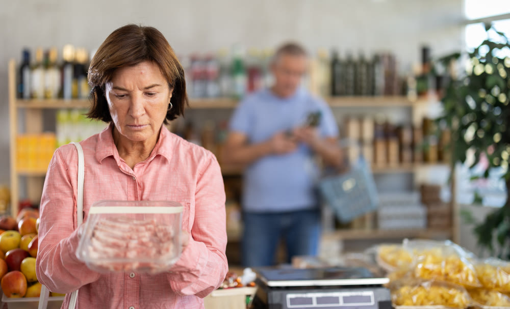 Senior woman choose raw pork meat for roasting in store, man client in background