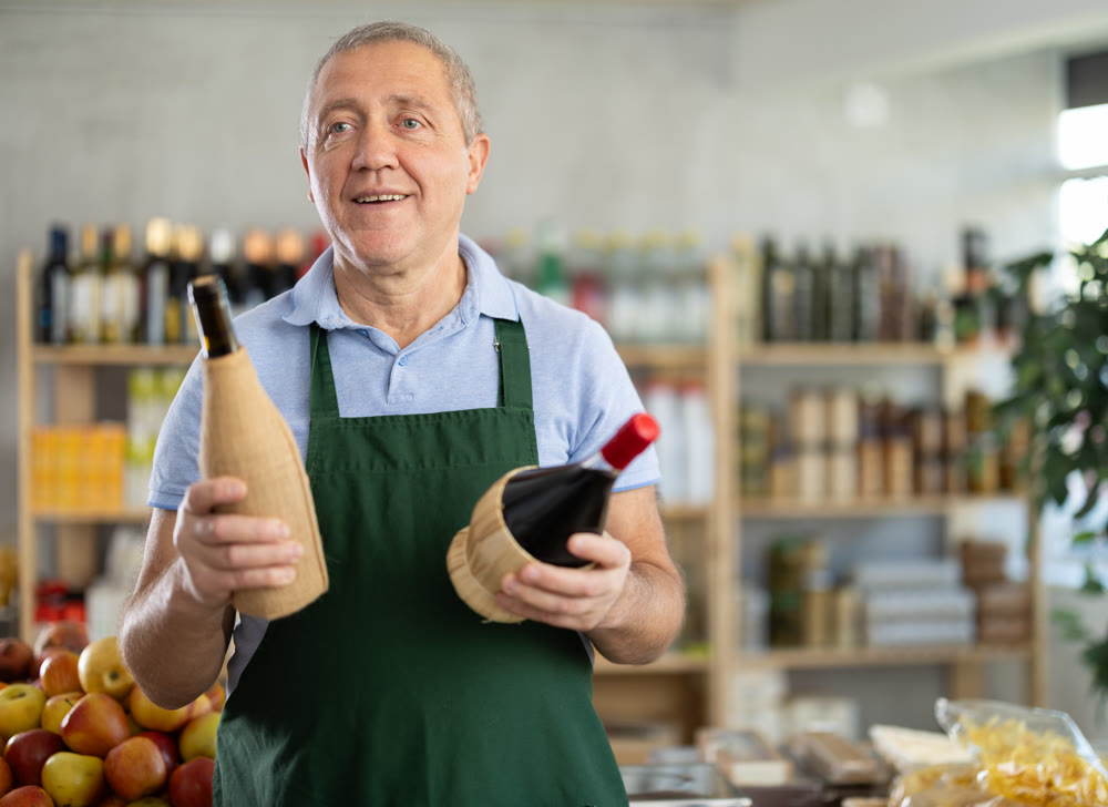 Senior man offers to buy alcohol, holds out bottles towards buyer, demonstrates best offers.