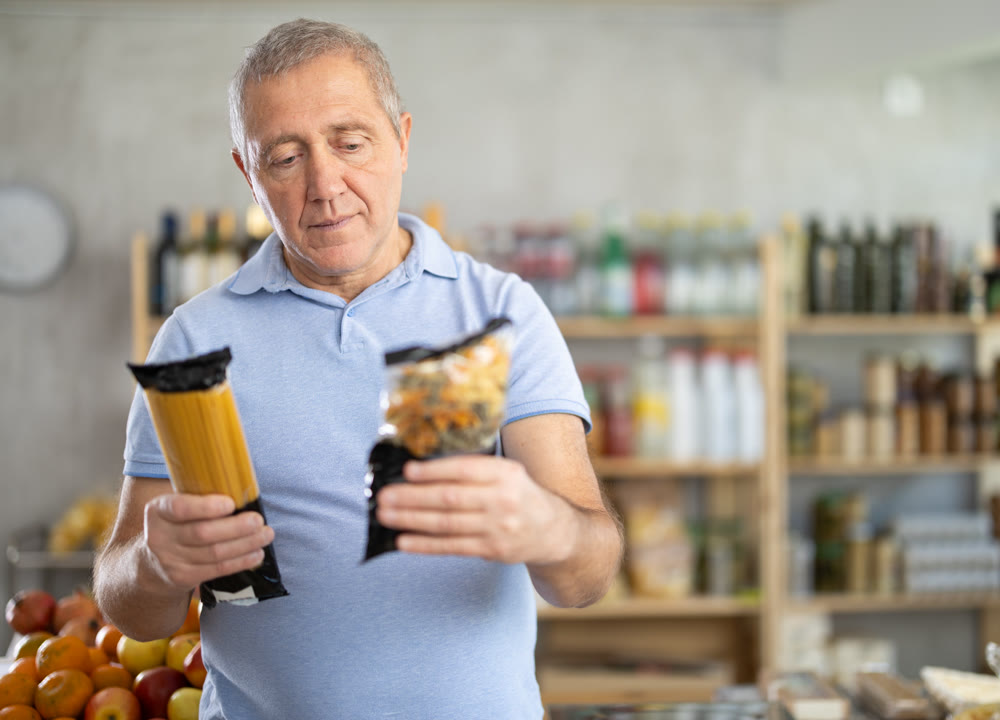 Senior man is holding two packages of noodles in hands, choosing food for garnish side dish
