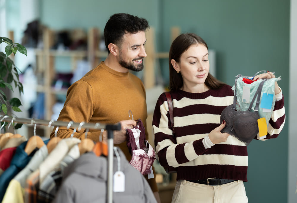 Happy couple dressing in ordinary clothes holding lingerie in hands and shopping in underwear store, man helping choose right size