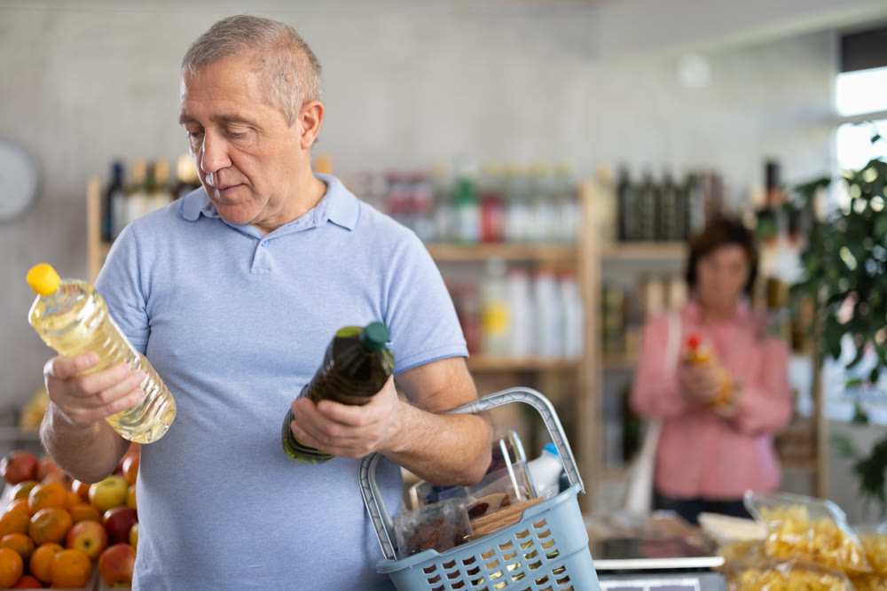Mature man buyer looks thoughtfully at bottles of vegetable oil, woman client in background