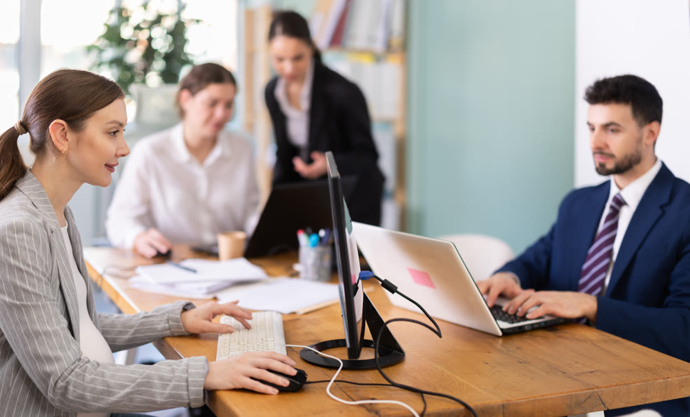 Pregnant woman is working on a laptop in an office while colleagues are discussing a project