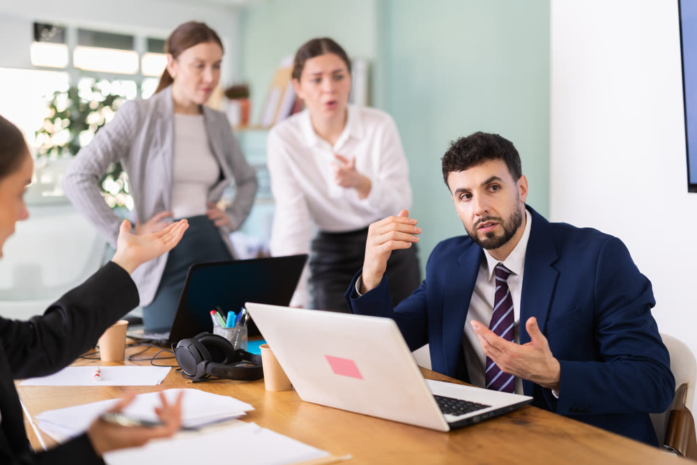 Group of people scolding sad male colleague sitting at laptop in office
