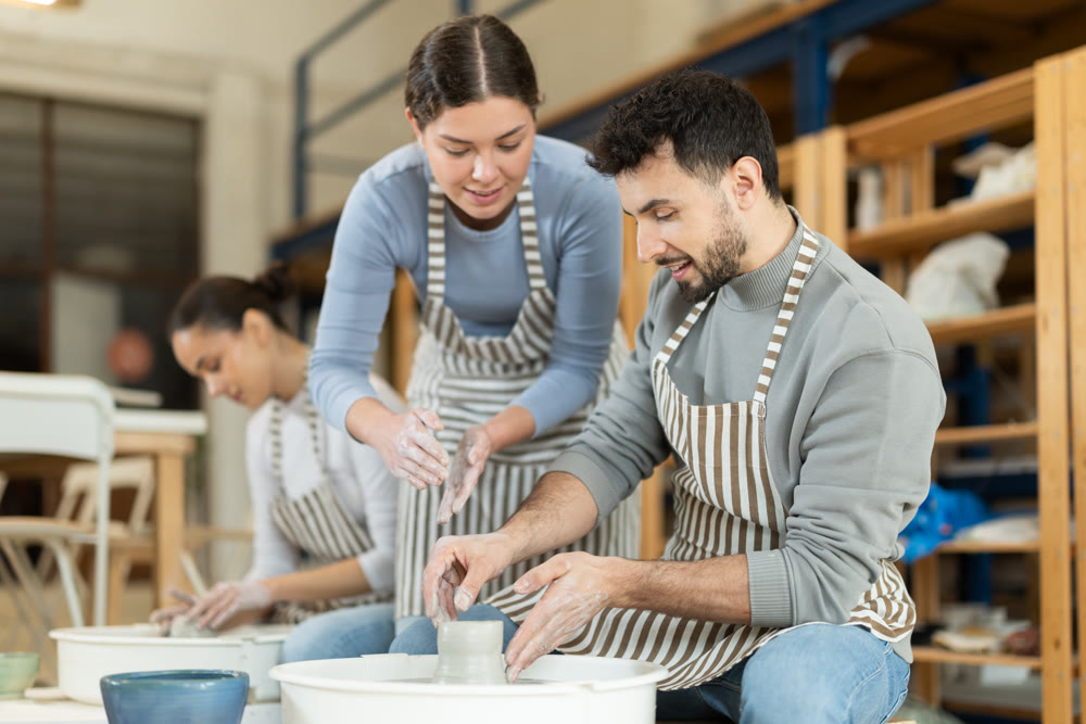 Female teacher help man sculpting ceramic product from clay on pottery wheel in workshop