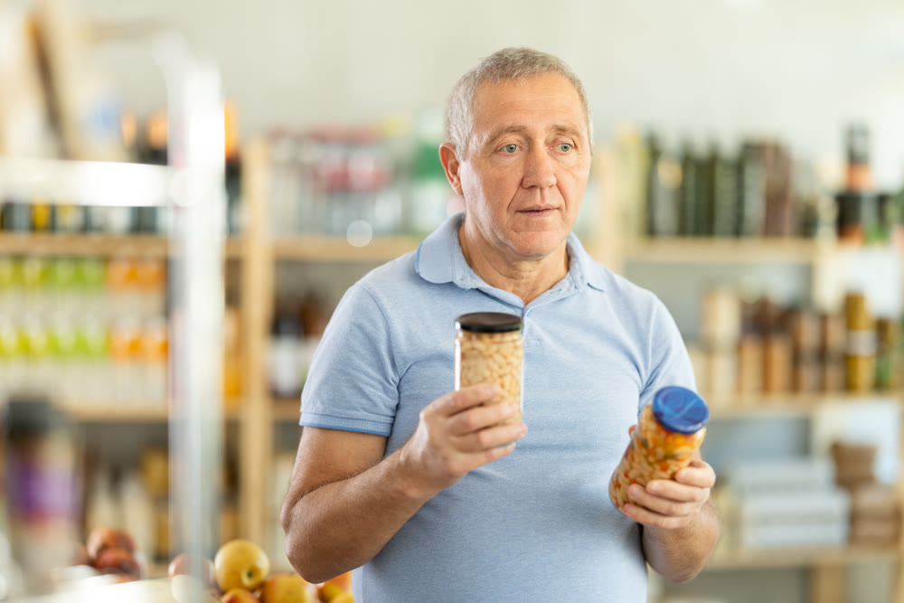 Senior man is holding two glass jars of preserves in hands, choosing pickled beans