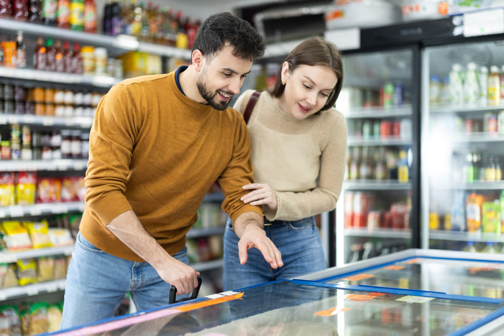 Positive young couple choosing cooled food product standing at fridge in grocery store