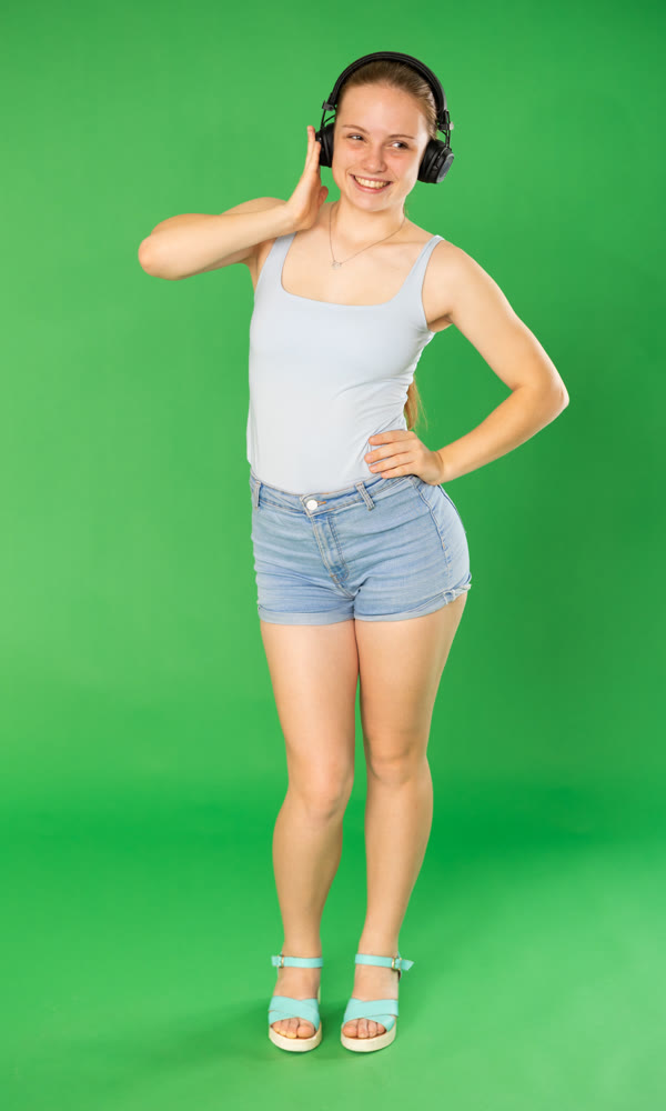 Passionate teenage girl in blue top listening and dancing to music on headphones on green background. Chroma key compositing
