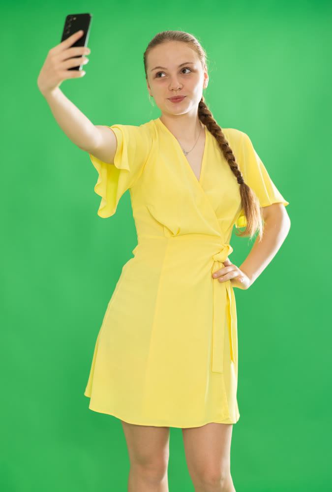 Smiling teenage girl in yellow dress takes selfie on green background. Chroma key compositing.