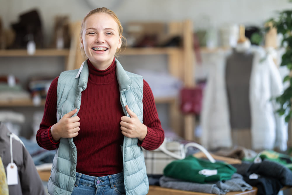 Positive teen girl trying on blue vest for cold season in clothing store