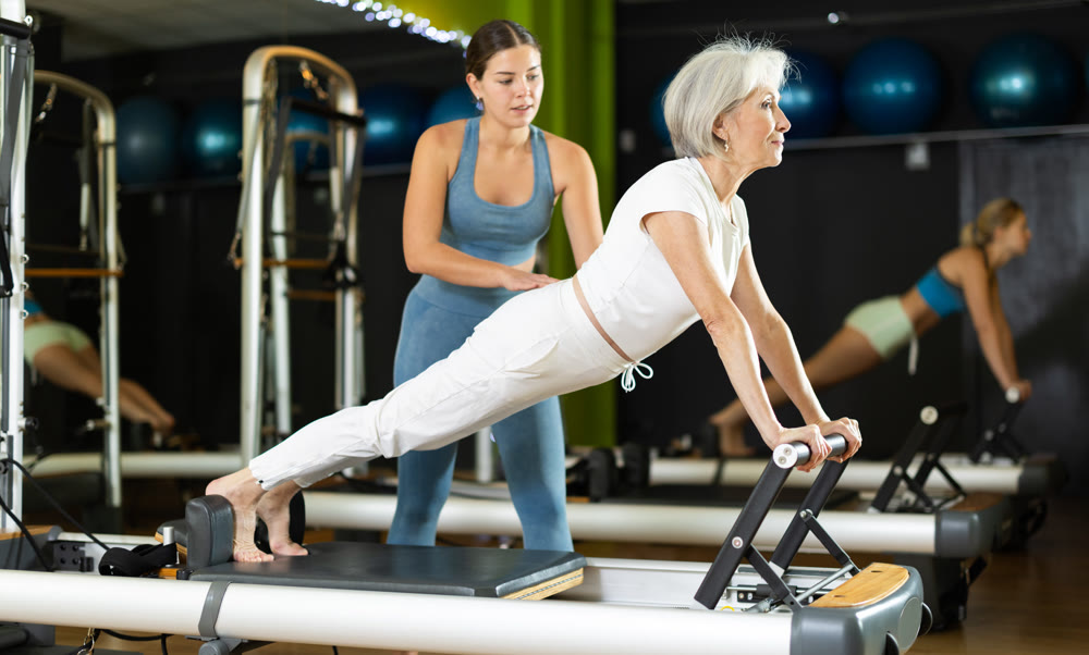 Girl trainer helps senior woman to perform plank exercises on pilates reformer