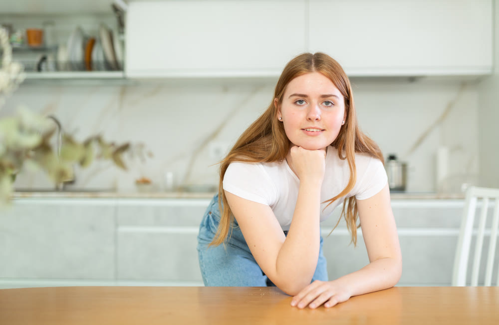 Portrait of positive teenage girl smiling at camera while leaning on countertop in cozy kitchen