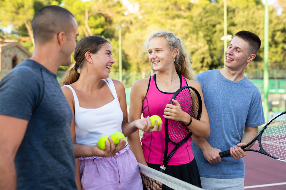 Group portrait of team of male and female partners tennis players standing and joyfully talking on tennis court at fitness club