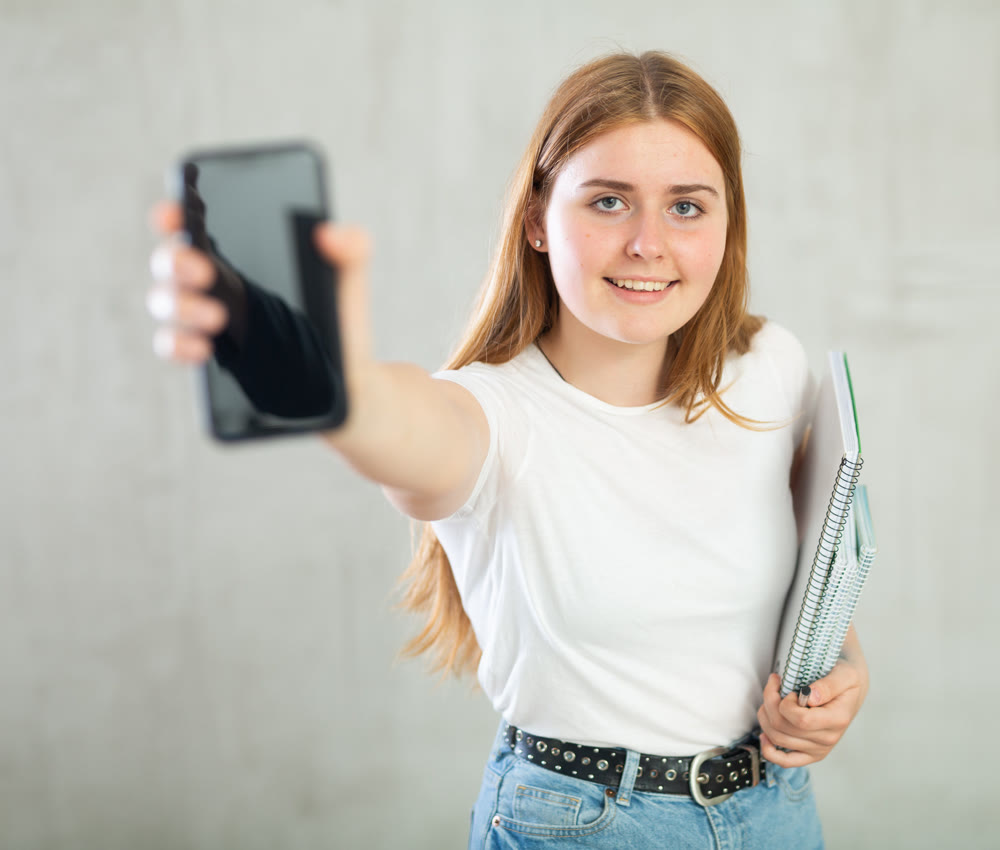 Happy teen girl, student in casual wear showing modern smartphone with empty black screen against gray wall background