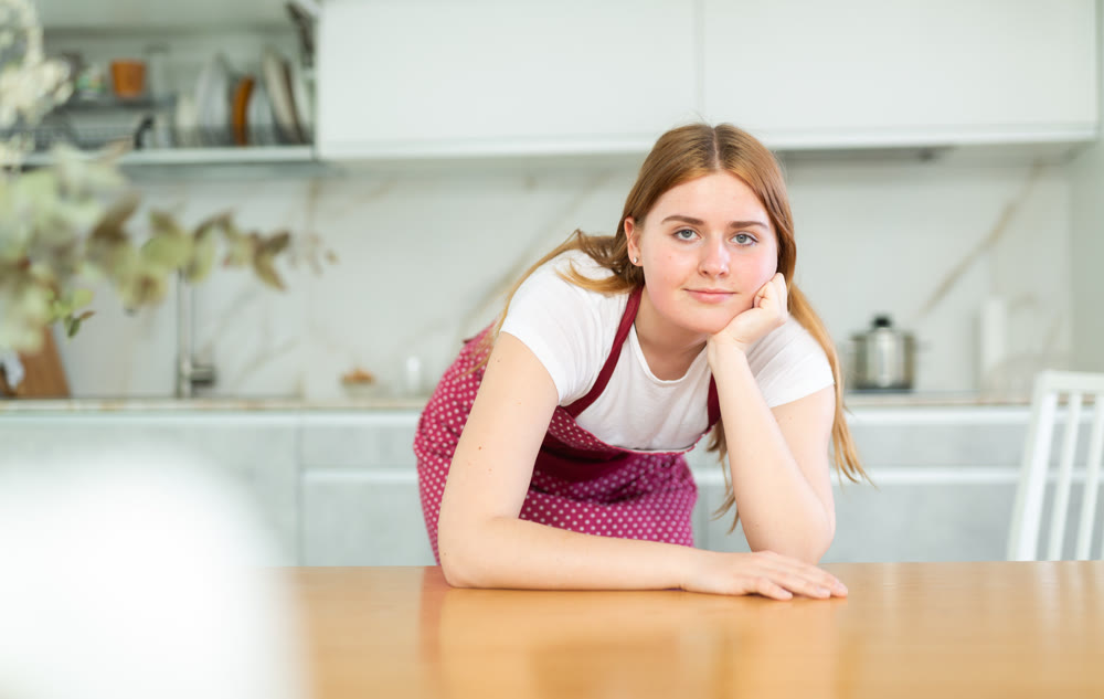 in kitchen,smiling young girl in red dotted apron props chin with fist and leans on kitchen table.