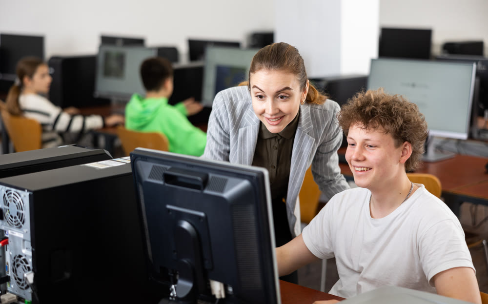 Smiling female teacher and teenage boy looking at monitor of PC