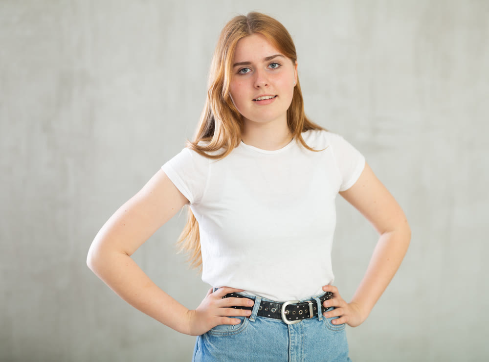 Portrait of positive teen girl in casual wear smiling at camera while posing against concrete gray wall background