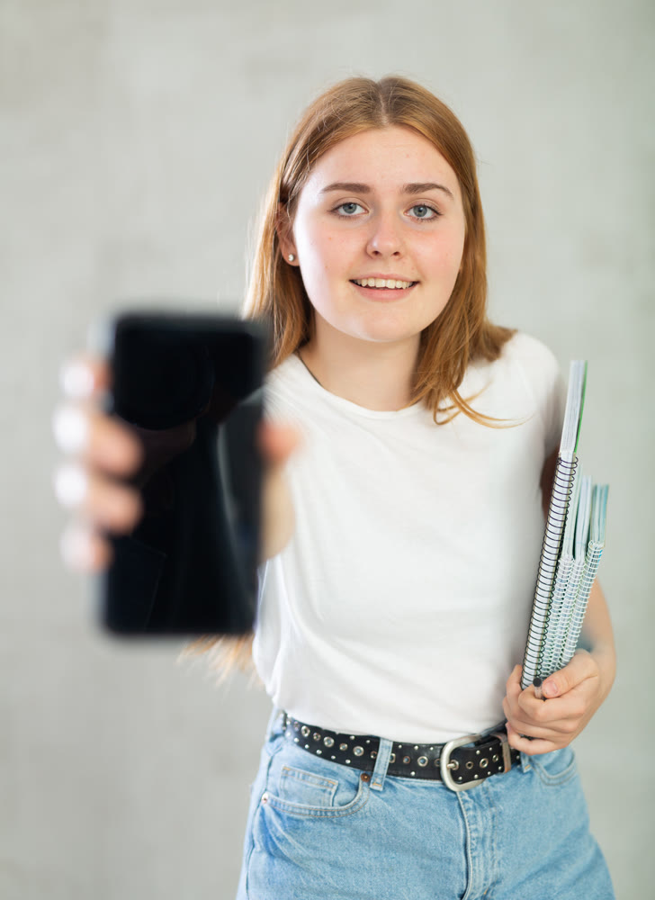 Smiling teenage girl, student with notebooks under her arm and mobile phone with blank black screen at gray wall background. Free copy space for advertisement